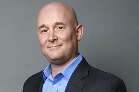 Bald man wearing a dark suit jacket and light blue collared shirt, smiling slightly, with a plain gray background.