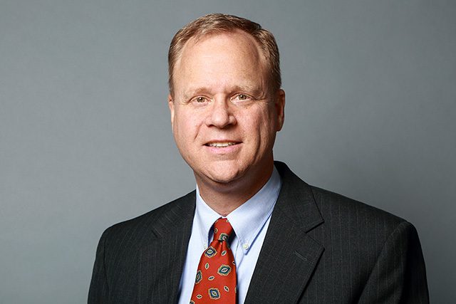 A middle-aged man in a dark suit, white shirt, and patterned red tie poses against a plain gray background.