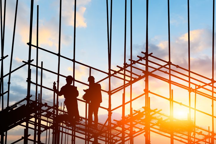 Two construction workers stand on scaffolding at a building site during sunset, with metal framework silhouetted against the sky.