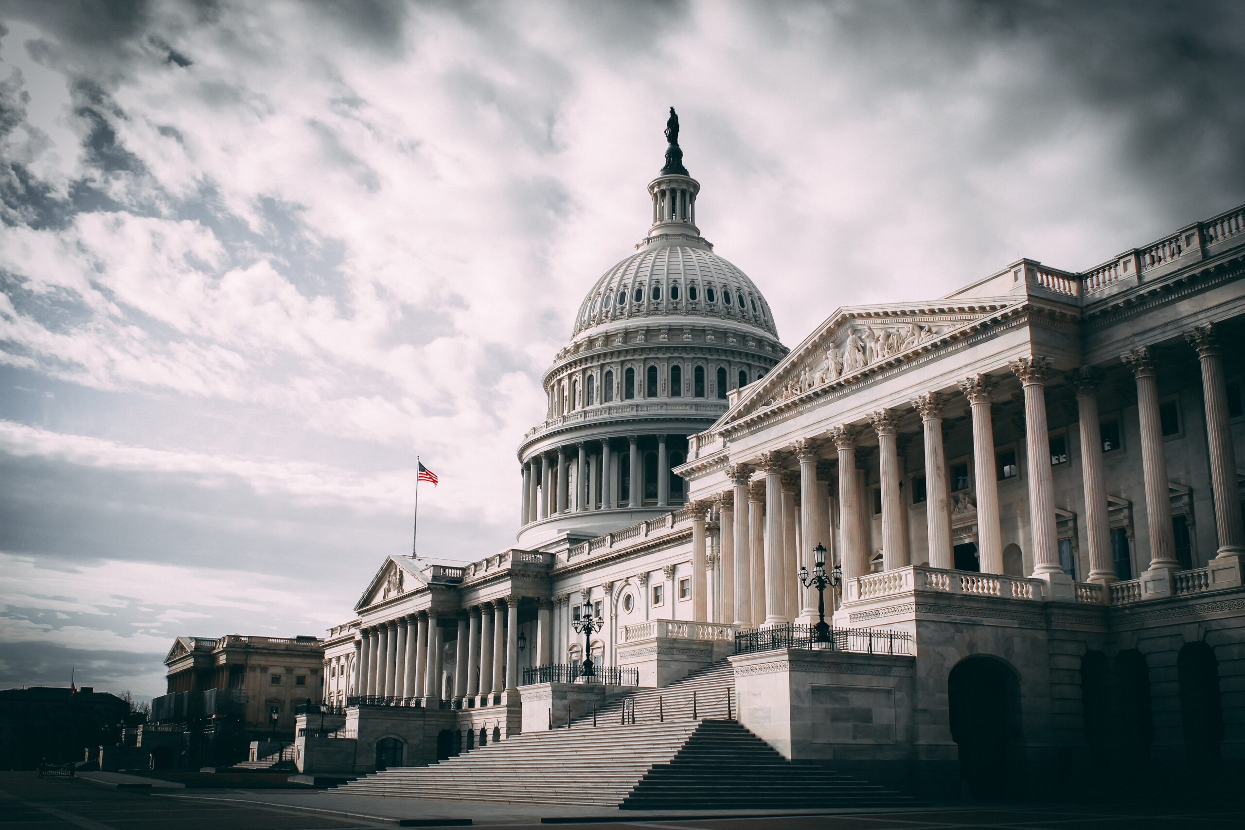 The United States Capitol building with an American flag, marble steps, and cloudy sky in the background.