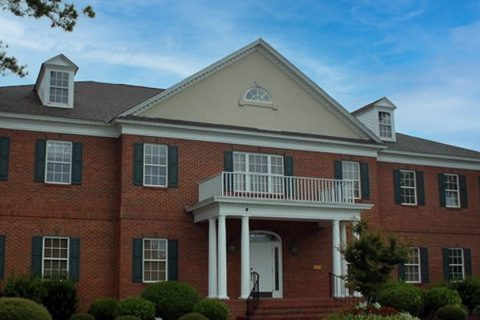A two-story red brick building with white columns, a balcony above the entrance, shuttered windows, and a gabled roof under a blue sky.