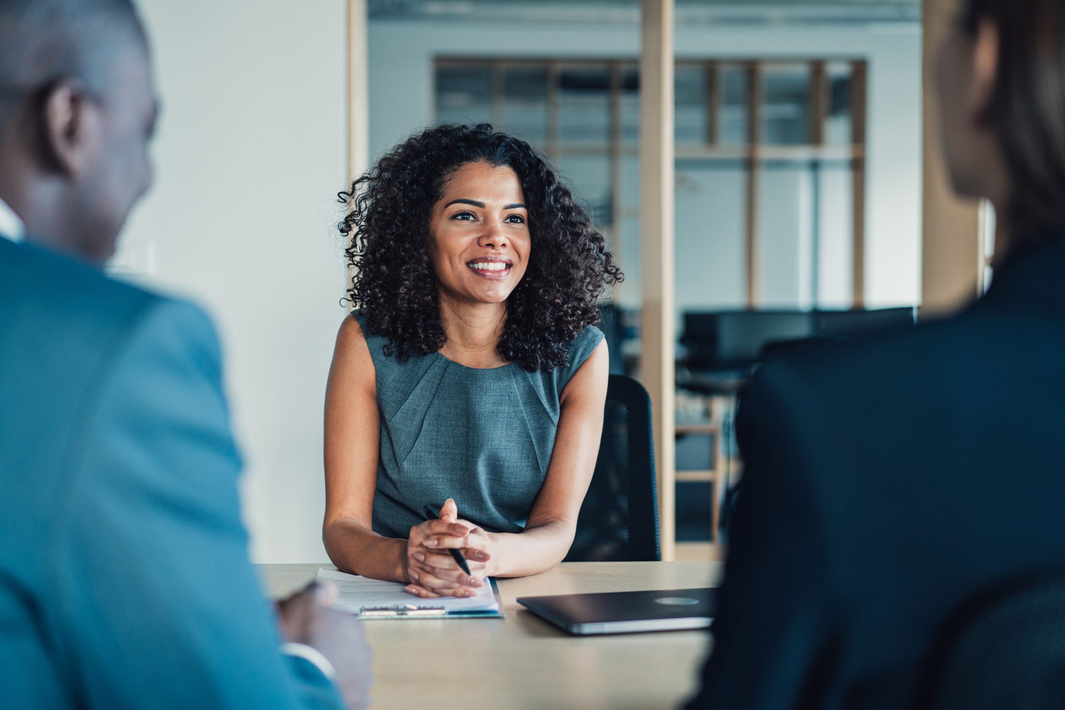 A woman in business attire sits at a conference table, smiling and speaking with two colleagues in an office setting.