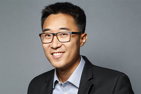 A man wearing glasses, a black blazer, and a light blue shirt smiles at the camera against a plain gray background.