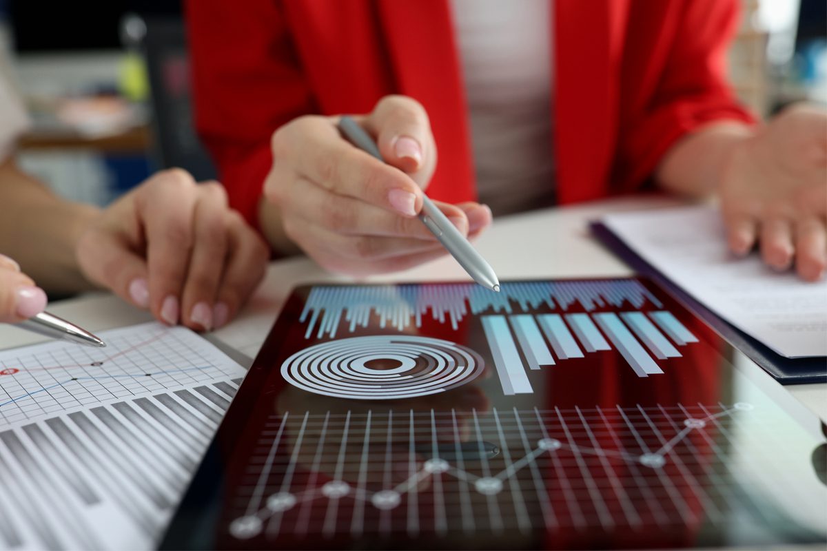Two people discussing financial graphs and charts displayed on a tablet and paper, with one person pointing at the tablet using a pen.