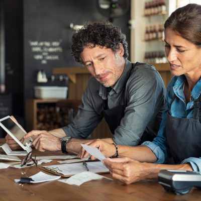 Two people wearing aprons sit at a table reviewing receipts and documents, with a tablet and calculator nearby in a cafe or small business setting.