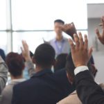 A group of people seated indoors raise their hands while facing a speaker standing in front of them.