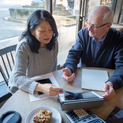 Two people sit at a table with papers, a tablet, and a calculator, discussing documents. A pastry and a coffee cup are also on the table.