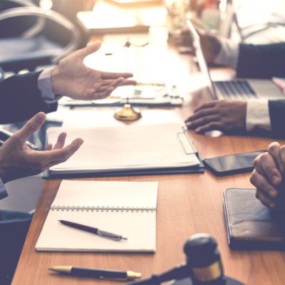 Two people sitting at a desk with open notebooks, a laptop, a gavel, and documents, engaged in a discussion or meeting.
