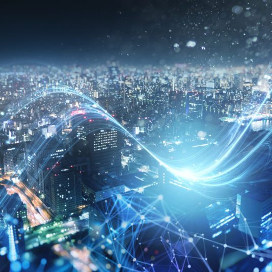 A cityscape at night with tall buildings and bright lights, overlaid with digital lines and network connections representing modern technology and connectivity.