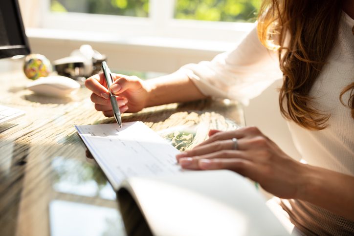 A person sits at a glass desk filling out a check with a pen, with sunlight coming through a window in the background.