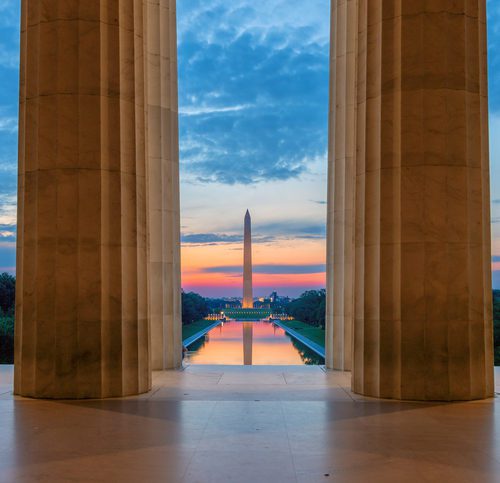 View of the Washington Monument and Reflecting Pool at sunset, framed by two large stone columns.