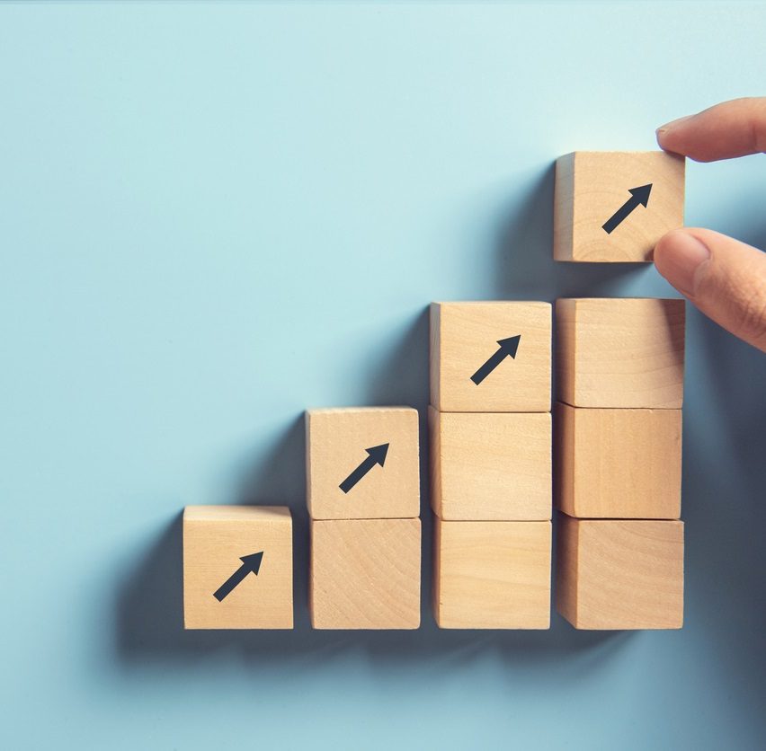 A hand stacks wooden blocks in ascending order, each block featuring an upward-pointing black arrow, against a light blue background.