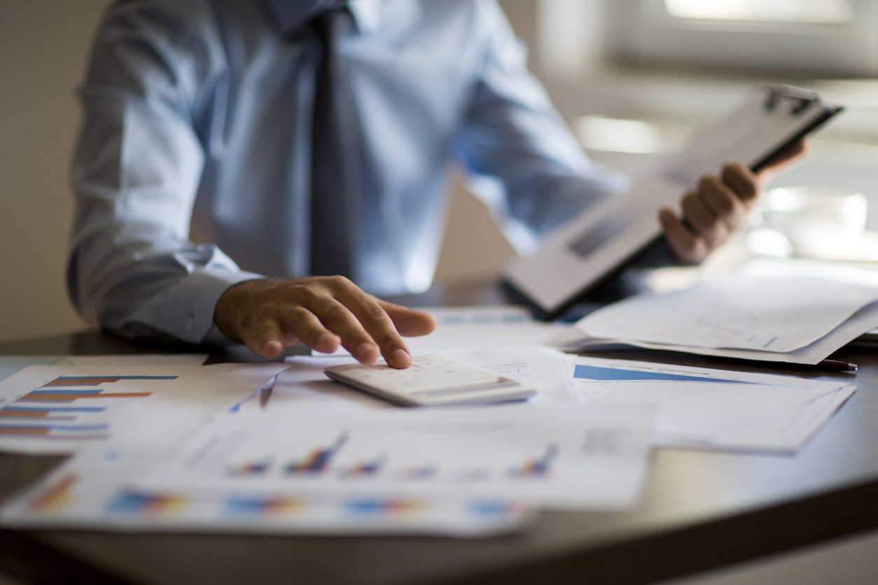 A person in business attire uses a calculator while reviewing documents and charts scattered across a desk.