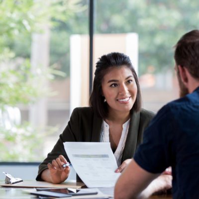 A woman holds a document and smiles while speaking to a man across a table in an office setting with large windows.