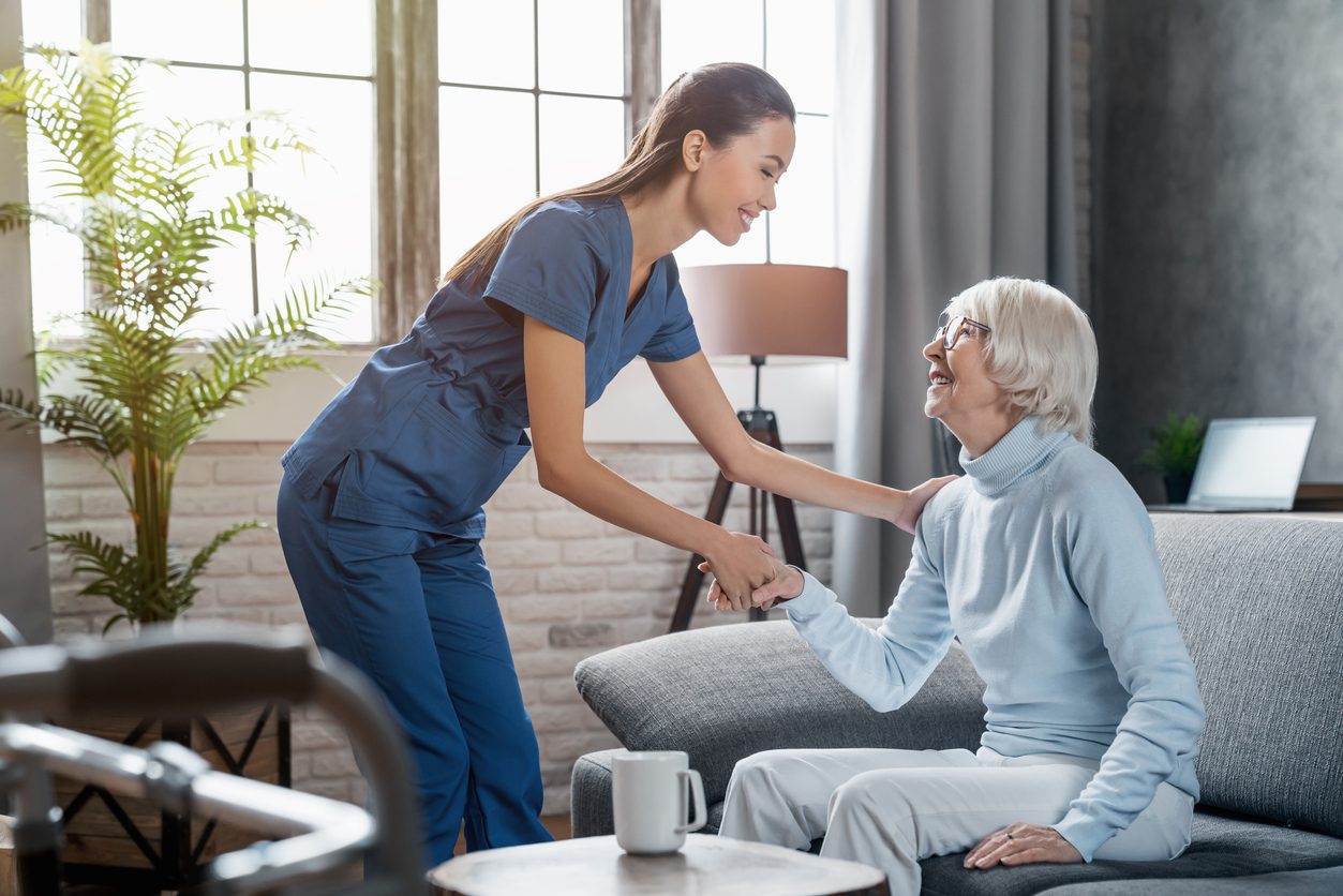 A nurse in blue scrubs helps an elderly woman sitting on a couch in a bright living room. The two women are smiling at each other.