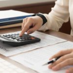 Person using a calculator and holding a pen, working with documents on a desk.