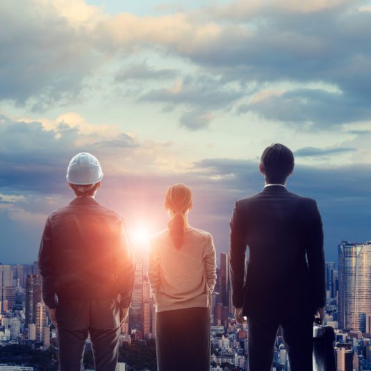 Three people, including a person in a suit, a person in a hard hat, and a person with a ponytail, stand facing a city skyline at sunset.
