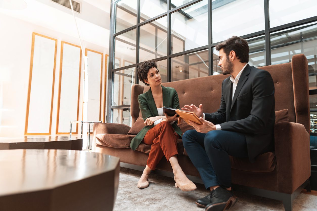 Two people in business attire sit on a brown sofa in a modern office, engaged in a serious conversation while holding documents.