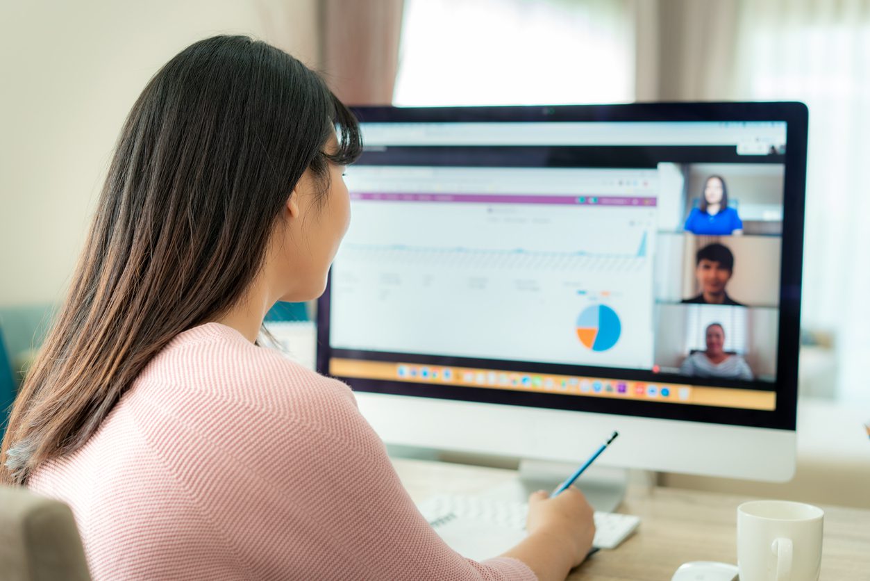 A woman sits at a desk viewing data charts on a computer screen while participating in a video conference with three people.