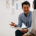 A man in a blue shirt is smiling and talking to a woman while holding papers in an office setting.