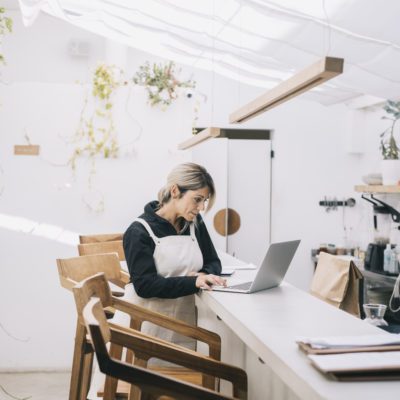 Person wearing an apron uses a laptop at a counter in a bright, modern café or kitchen with wooden chairs and plants on the wall.