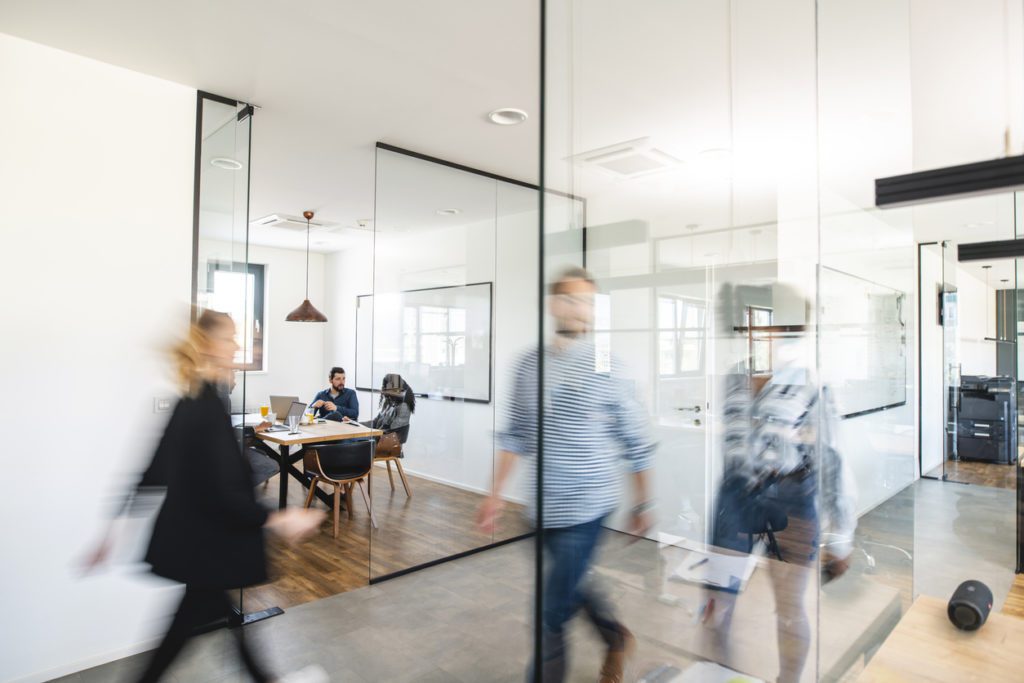 Modern office with people walking in a hallway and two individuals seated at a table in a glass-walled meeting room. The scene appears busy and bright.