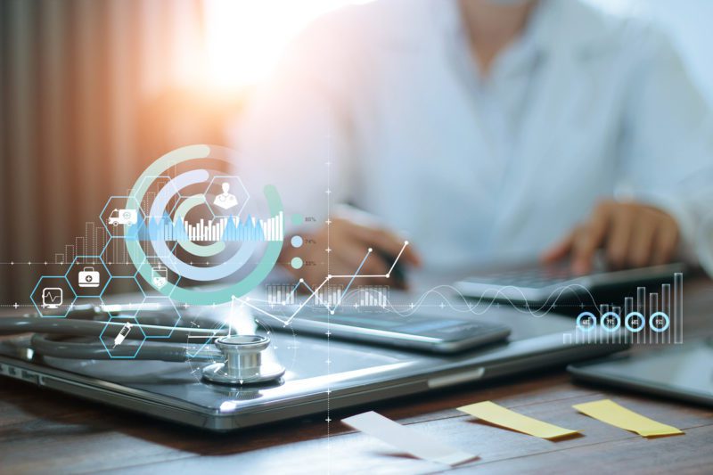 A stethoscope, clipboard, and smartphone on a desk with digital medical graphics overlay; a person in a white coat works in the background.