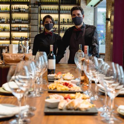 Two restaurant staff in black uniforms and face masks stand behind a set table with wine glasses, wine bottles, and platters of food in a modern dining area.