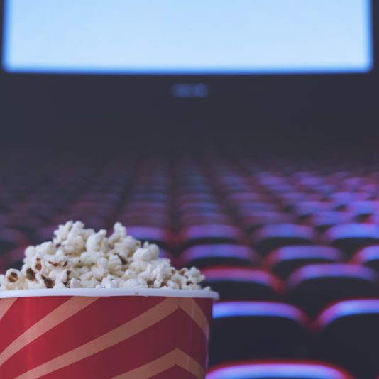 A striped tub of popcorn in the foreground with empty red theater seats and a blank movie screen in the background.