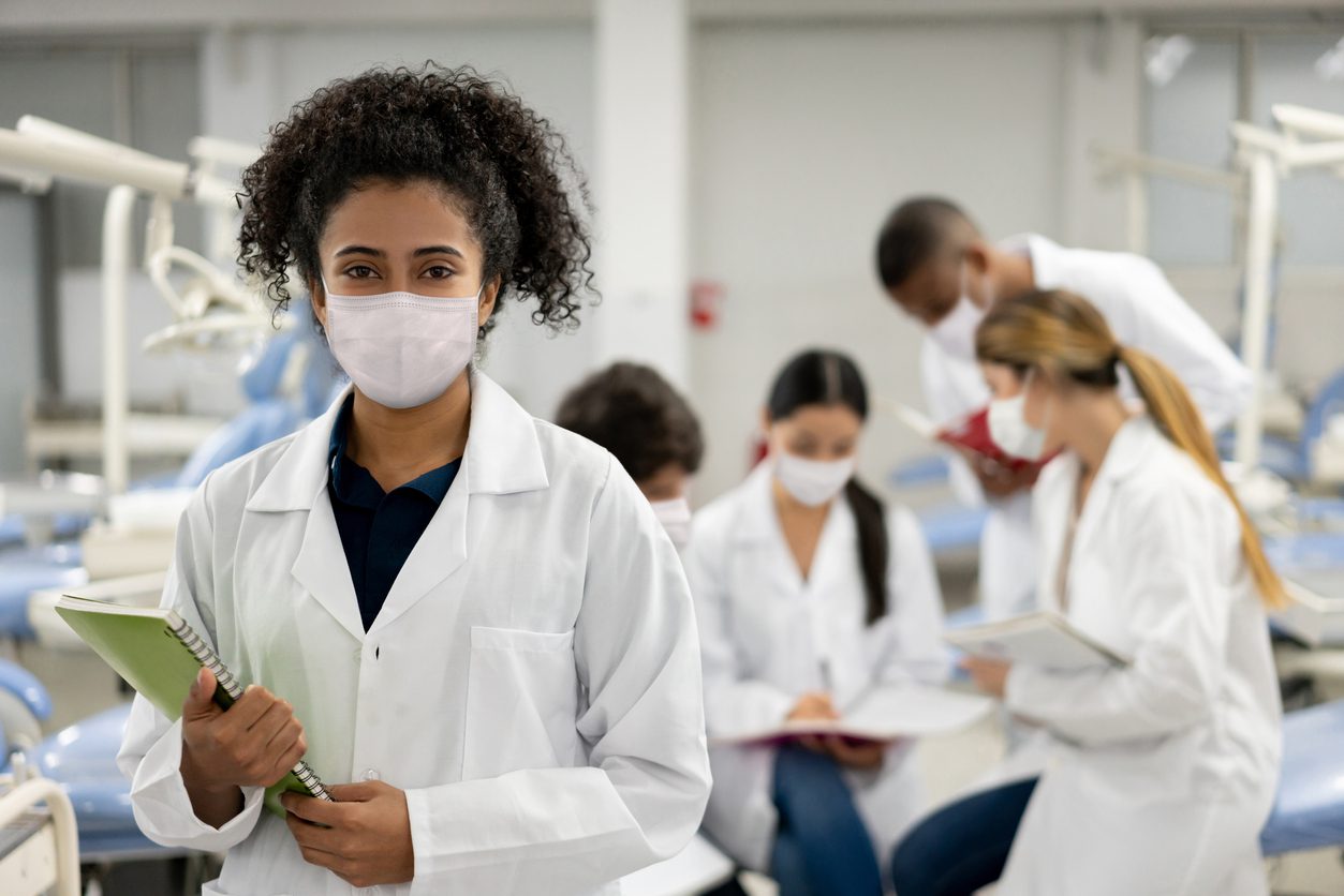 A woman in a lab coat and mask holds a notebook in a dental clinic, while four other masked people in lab coats work together in the background.