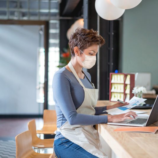 A woman wearing a face mask and apron sits at a counter, working on a laptop and holding documents in a modern indoor setting.