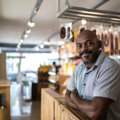 A man with a shaved head and beard stands behind the counter in a shop, smiling with arms crossed. Shelves and hanging products are visible in the background.