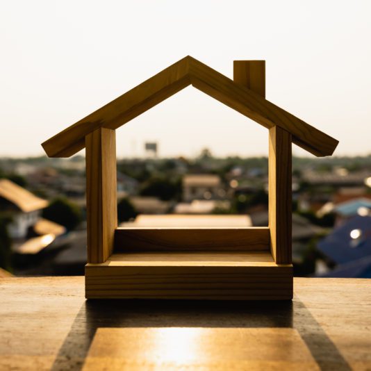A wooden model of a house is placed on a table, with a blurred view of houses and buildings in the background under sunlight.
