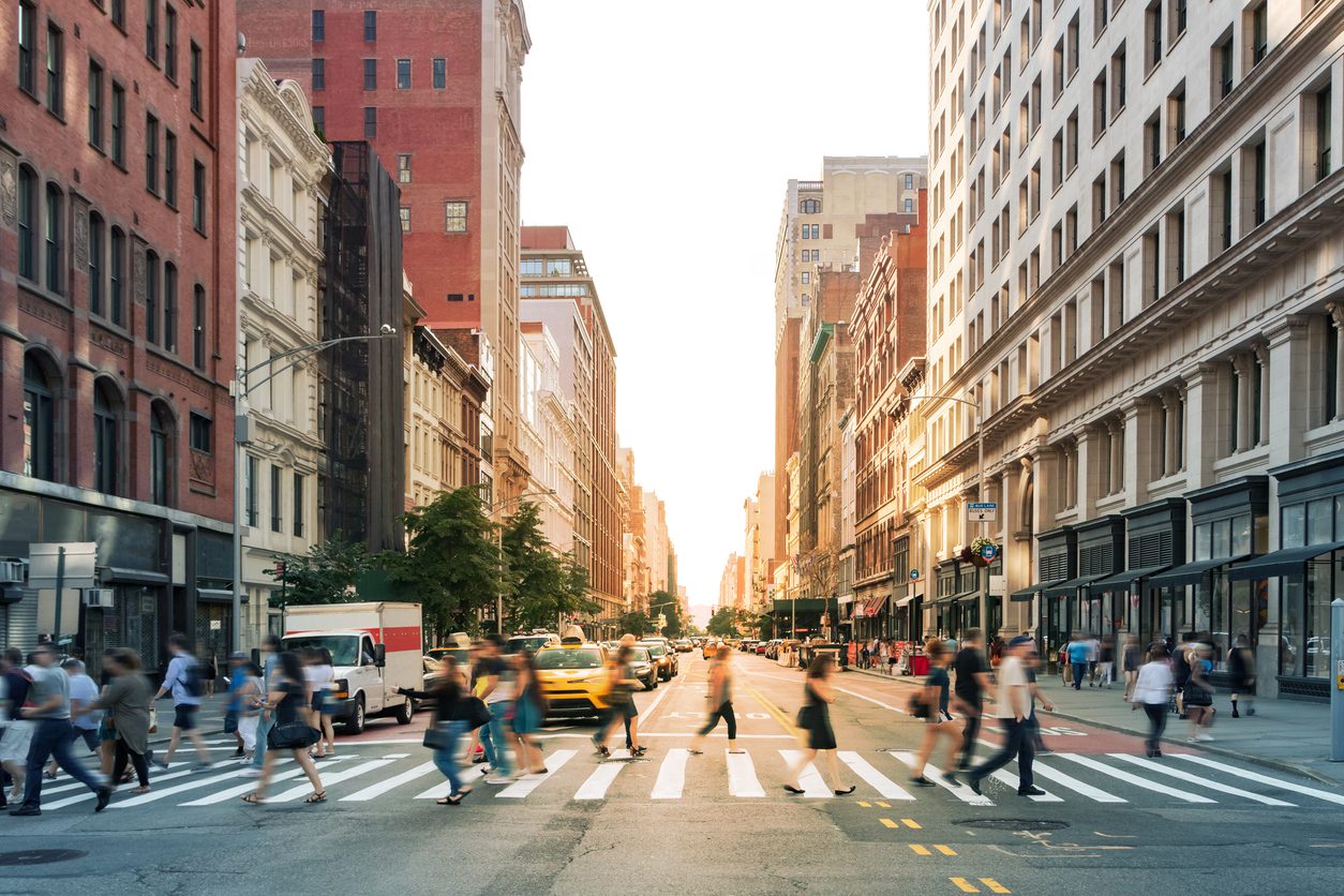 People crossing a busy city street at a crosswalk, with cars, taxis, and tall buildings lining both sides of the road at sunset.