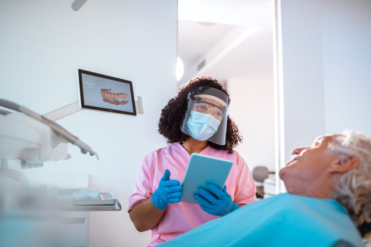 A dental professional in PPE holds a tablet while talking to a patient in a dental chair, with dental images displayed on a monitor nearby.