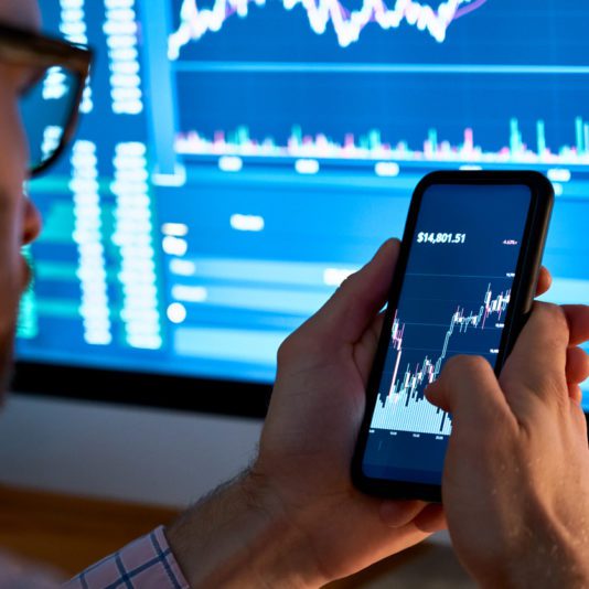 A person holding a smartphone displaying a stock market chart, with a large computer screen showing financial graphs in the background.