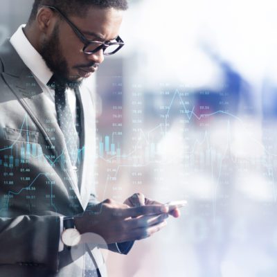 A man in a suit looks at his smartphone with financial charts and stock market data superimposed in the background.