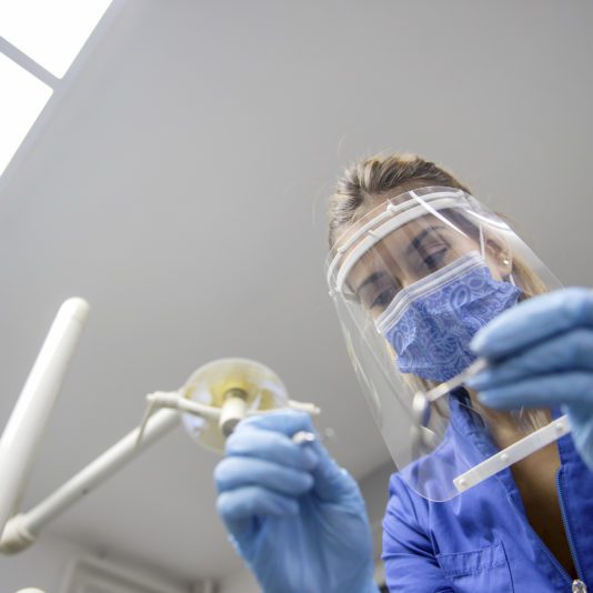 A dental professional wearing a face shield, mask, and gloves examines dental tools in a clinic setting.