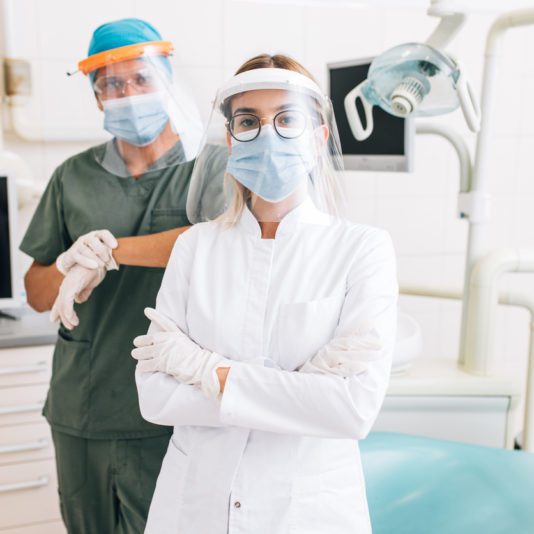 Two dental professionals wearing face masks, gloves, and face shields stand in a dental clinic with equipment in the background.
