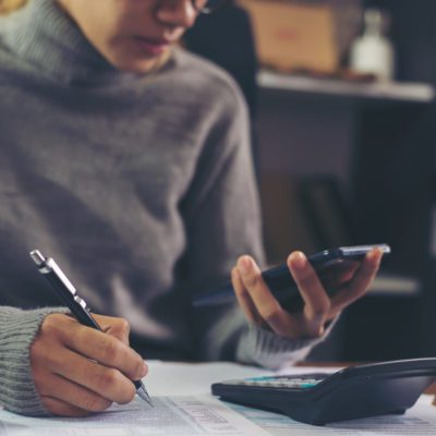 Person wearing a gray sweater using a pen to write on documents while holding a smartphone, with a calculator on the desk.