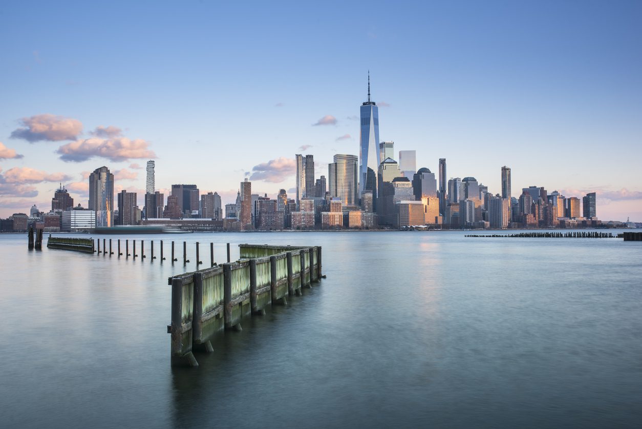 View of the Manhattan skyline at sunset with the One World Trade Center visible and a wooden pier extending into the Hudson River in the foreground.