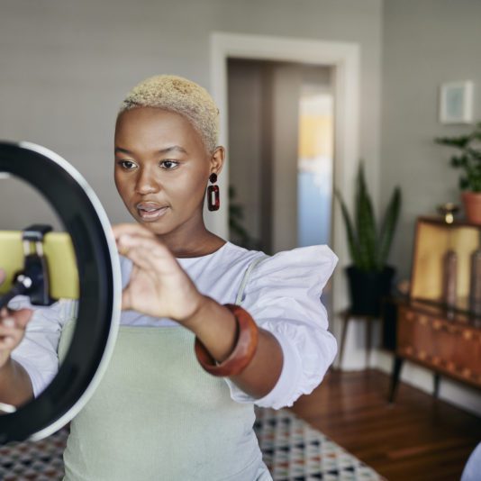 A person adjusts a smartphone on a ring light stand in a living room with plants and wooden furniture in the background.