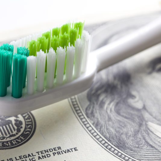 A toothbrush with green, blue, and white bristles rests on top of a U.S. dollar bill.