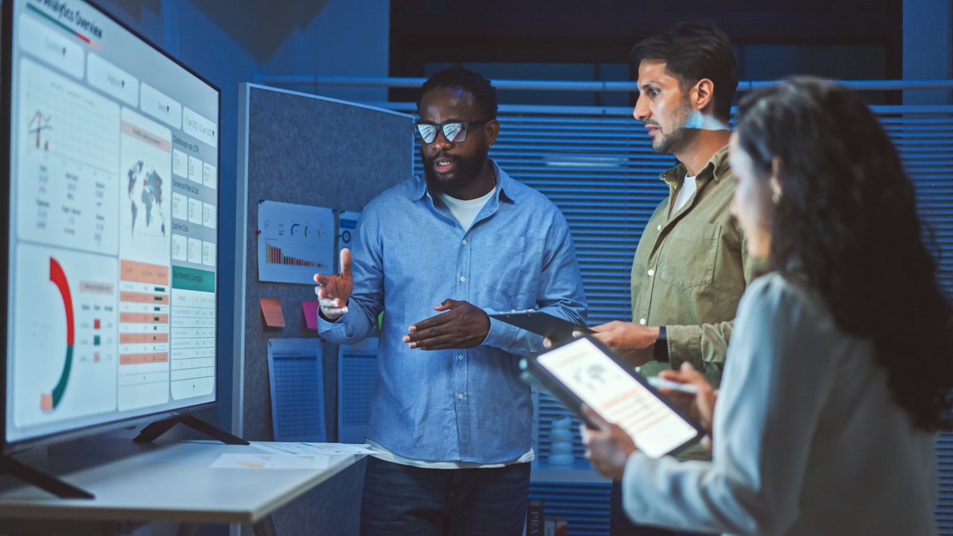 Three people in an office discuss data displayed on a large screen showing charts, graphs, and statistics while holding digital tablets and documents.