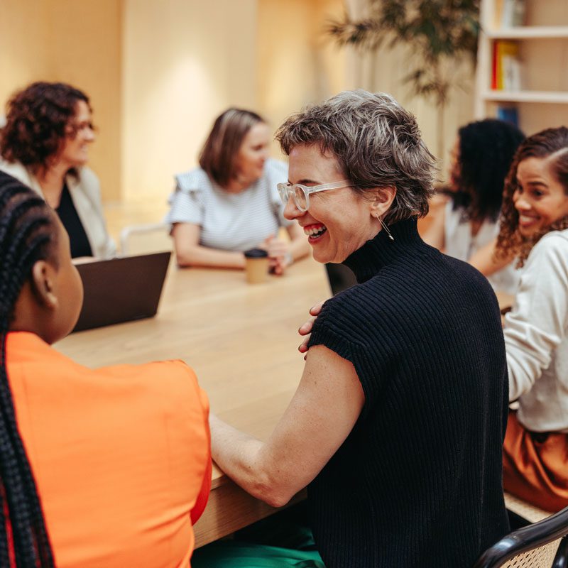 A group of people sit around a conference table engaged in discussion, with one woman in the foreground smiling and talking to another attendee.