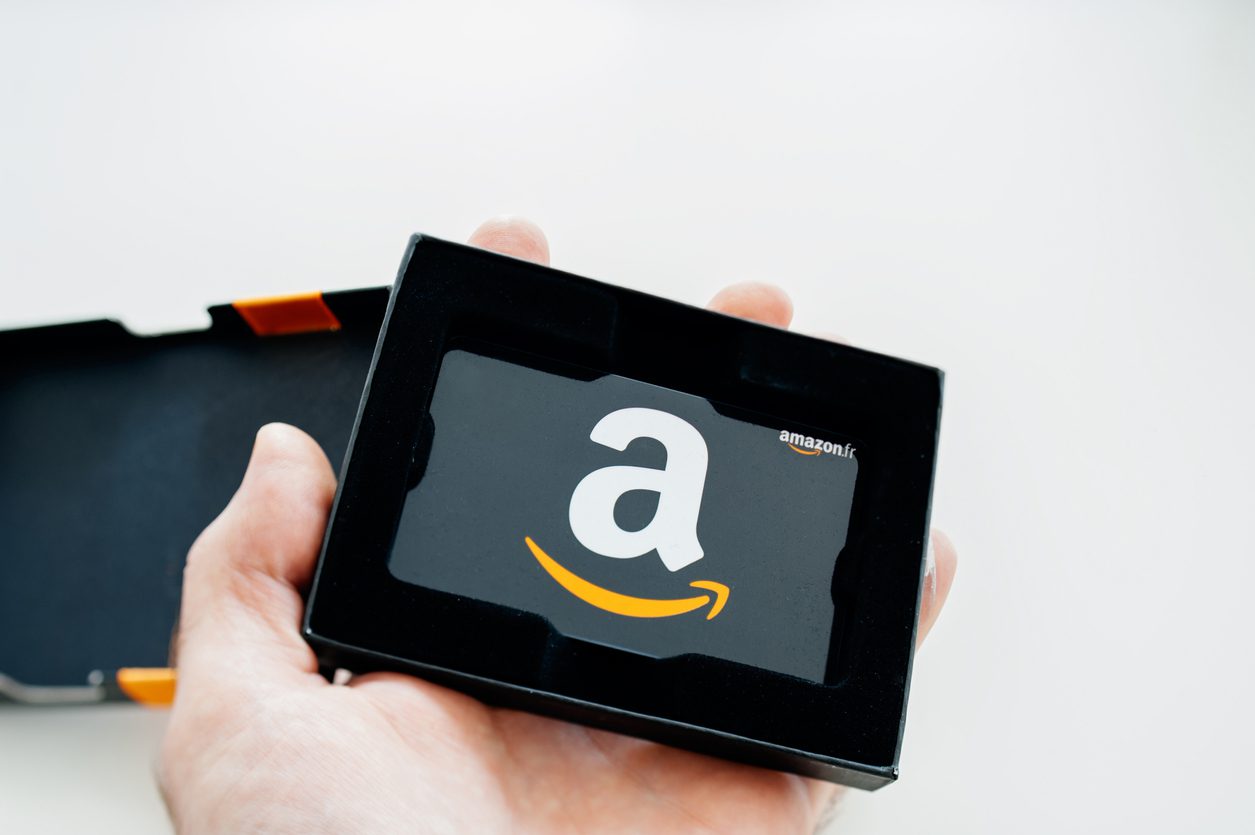 A hand holds an Amazon gift card in black packaging, with the large white Amazon “a” logo and yellow arrow visible.