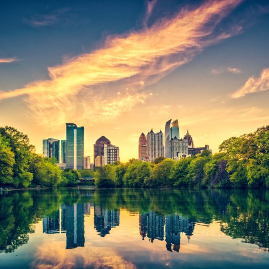 Atlanta city skyline at sunset, tall buildings reflected in a calm lake, with green trees along the water’s edge and vibrant clouds in the sky.