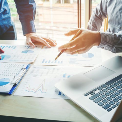 Two people reviewing graphs and charts on paper at a desk with a laptop, discussing financial or business data in a bright office setting.