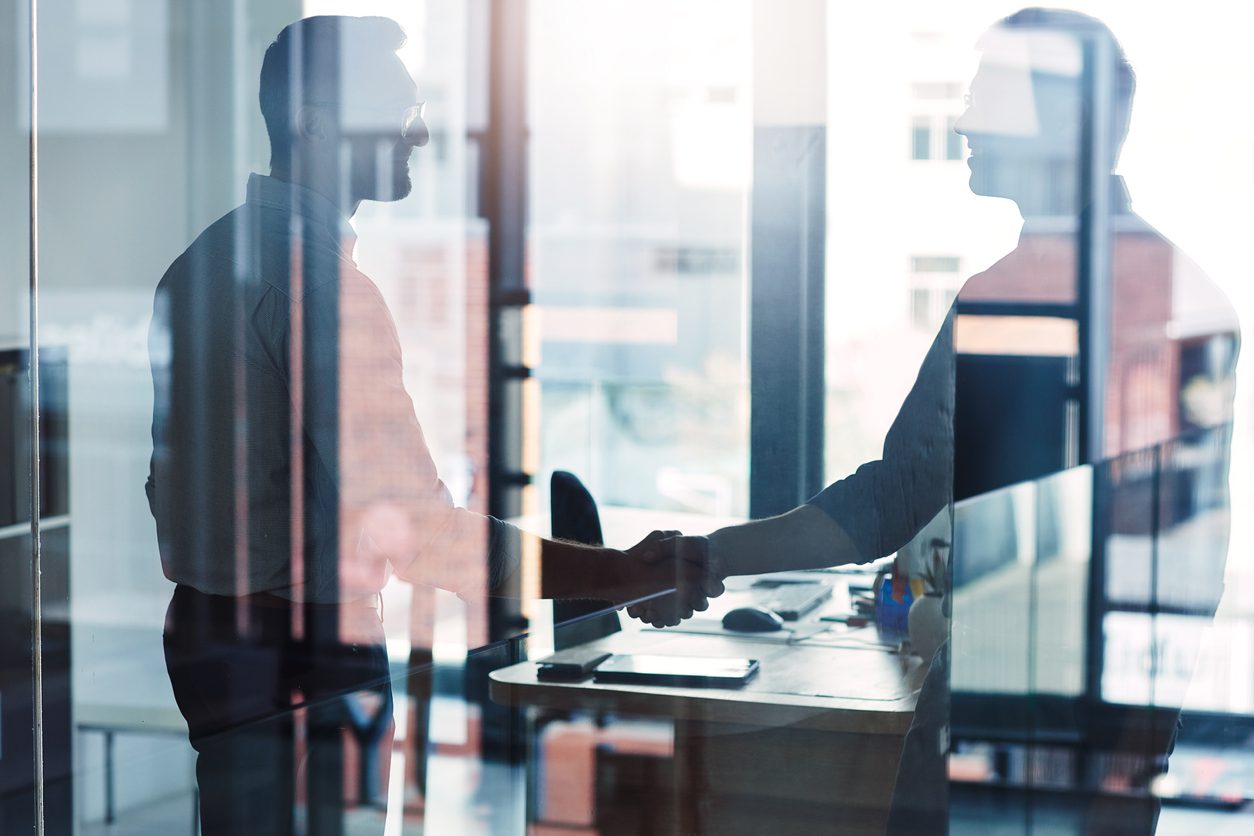 Two men are shaking hands in an office, seen through a glass wall with reflections.
