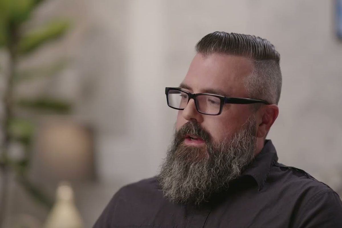 A man with glasses, a styled beard, and short-cropped hair sits indoors, wearing a dark shirt with a blurred background behind him.